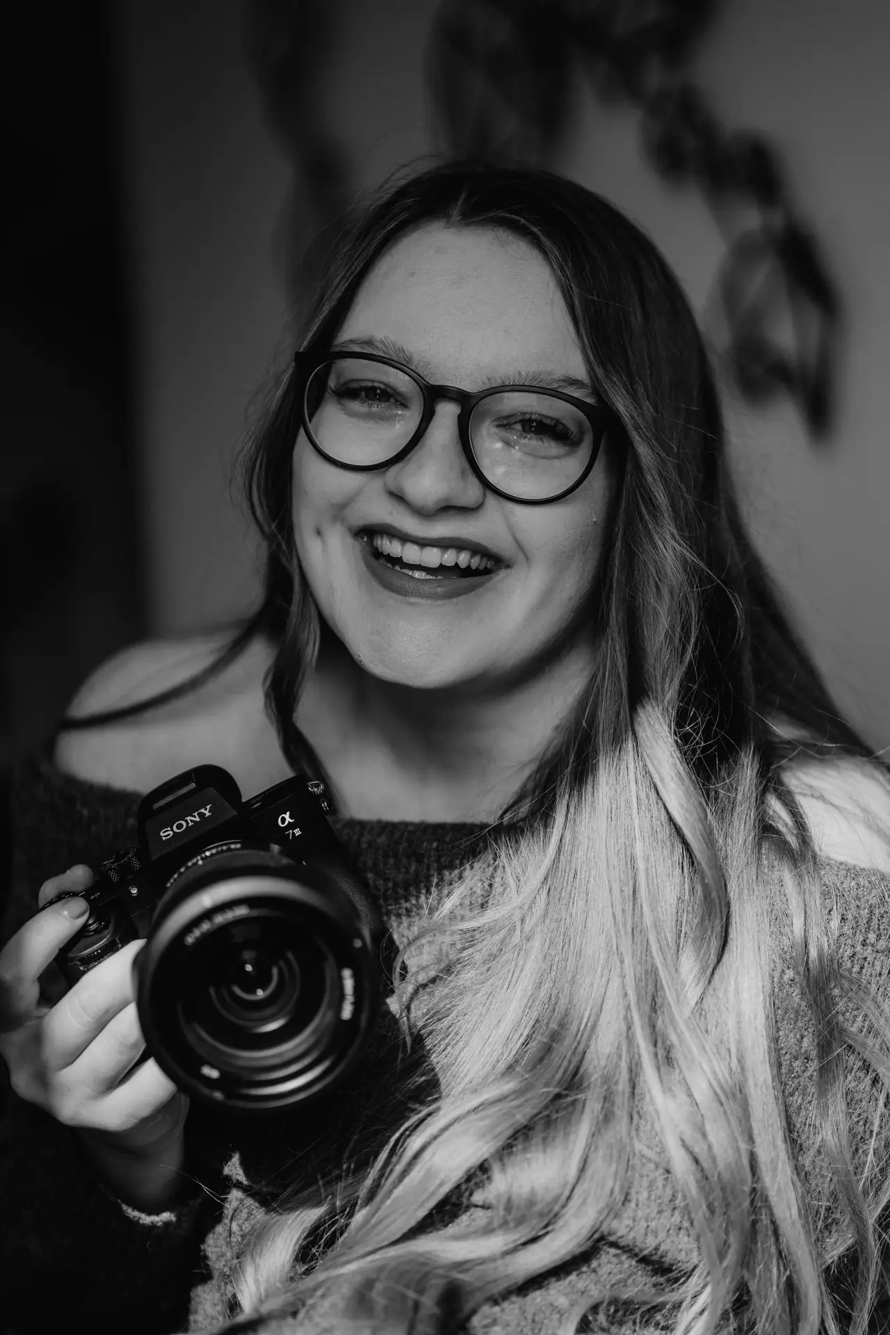 Eine schwarz-weiß Aufnahme einer Frau mit langem Haar und Brille die lächelt und eine Sony-Kamera in der Hand hält.