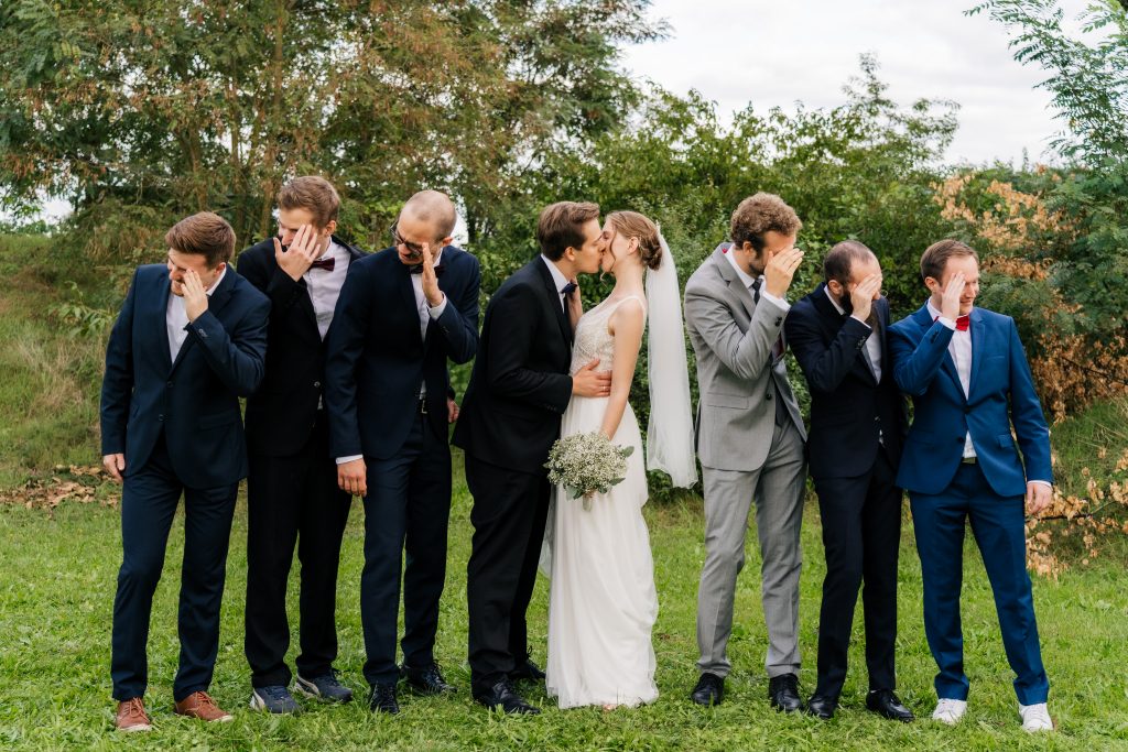 Gruppenfoto in der Reihe im Grünen draußen, auf dem sich das Brautpaar küsst und alle Junggesellen sich die Augen zuhalten und wegschauen