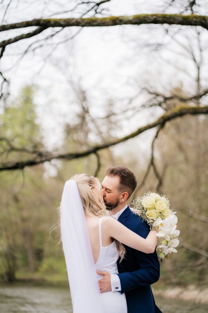 Braut mit langem Schleier und weißem Blumenstrauß küsst sich innig mit ihrem Ehepartner in einem blauen Anzug in einem Park mit See im Hintergrund