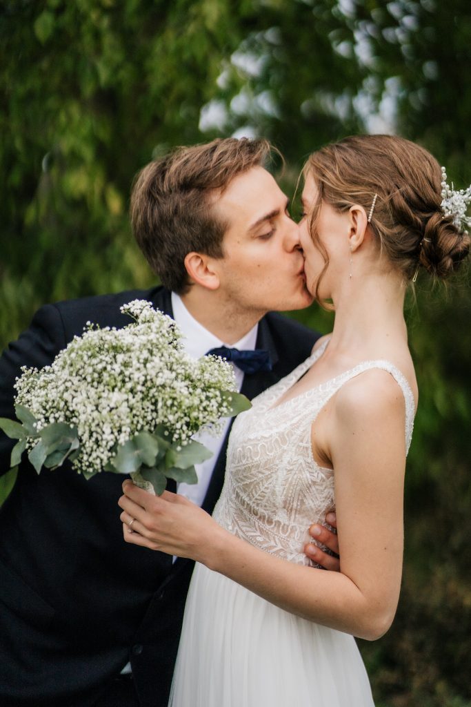ein Bräutigam küsst seine Braut die einen weißen Blumenstrauß hält und ein Boho Hochzeitskleid trägt überschwänglich