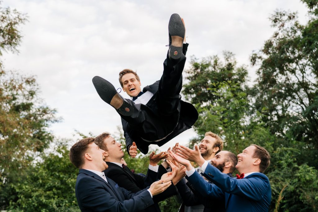 ein Mann wird von einer Gruppe von Männer in die Luft geschleudert und lacht dabei in die Kamera.