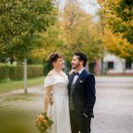 Eine Braut in einem Brautkleid hält einen Blumenstrauß und der Bräutigam in Anzug stehen zwischen herbstlichen Bäumen und umarmen sich seitlich an der Hüfte