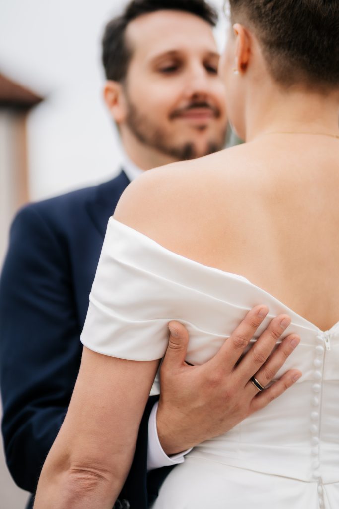 Der Bräutigam umarmt die Braut von vorne und hält sie am Rücken fest, ihr Kleid hat eine Knopfleiste und ist halb rückenfrei.